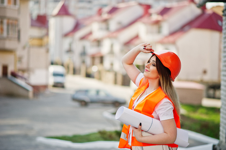 Engineer builder woman in uniform waistcoat and orange protective helmet hold business drawing paper roll against new building. Property living block theme.の写真素材