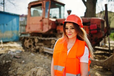 Engineer builder woman in uniform waistcoat and orange protective helmet against bulldozer.の写真素材