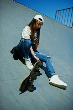 Young teenage urban girl with skateboard, wear on glasses, cap and ripped jeans at skate park on the evening.の写真素材