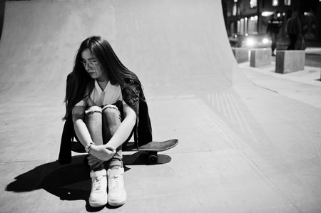 Young teenage urban girl with skateboard, wear on glasses, cap and ripped jeans at skate park on the evening.の写真素材