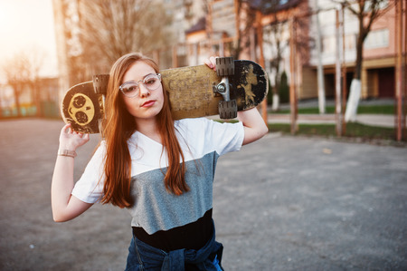 Young teenage urban girl with skateboard, wear on glasses, cap and ripped jeans at the yard sports ground on sunset.の写真素材