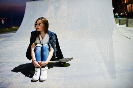 Young teenage urban girl with skateboard, wear on glasses, cap and ripped jeans at skate park on the evening.の写真素材