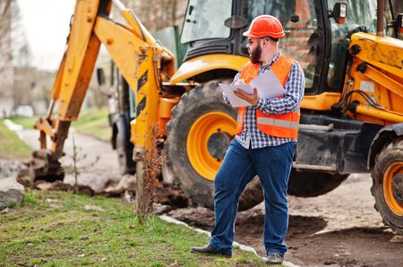 Brutal beard worker man suit construction worker in safety orange helmet, sunglasses against traktor...