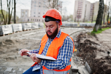 Brutal beard smoking worker man suit construction worker in safety orange helmet sitting on pavement, break at work, and read working notebook entries.の写真素材