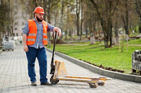Brutal beard worker man suit construction worker in safety orange helmet with pallet truck.の写真素材