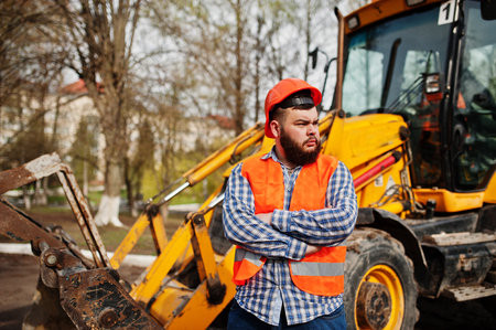 Brutal beard worker man suit construction worker in safety orange helmet against traktor.