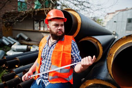 Brutal beard worker man suit construction worker in safety orange helmet near steel pipes with tape measure at hand.の写真素材