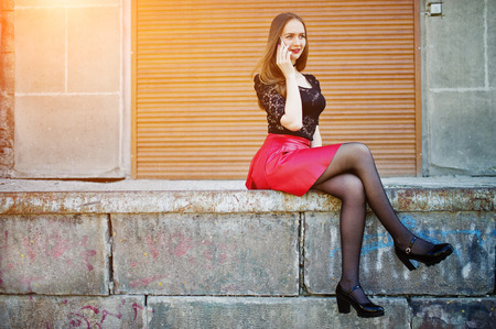 Portrait of girl with black choker on her neck, red leather skirt and mobile phone at hand against orange shutter.の写真素材