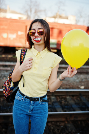 Young teenage girl standing on the platform at the train station with funny lips on stick and balloon at hand, wear on yellow t-shirt, jeans and sunglasses, with backpack.の写真素材