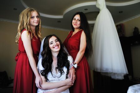 Happy cheerful brunette bride wearing at her room with two bridesmaids on red dress.の写真素材