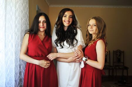 Happy cheerful brunette bride wearing at her room with two bridesmaids on red dress.の写真素材
