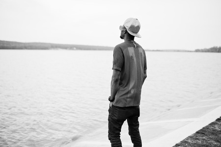 Stylish african american boy wear at cap, football t-shirt and sunglasses stay against lake beach. Black sports man portrait.の写真素材