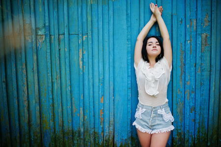 Portrait of sexy brunette girl on women's jeans shorts and white blouse against blue wooden background.の写真素材