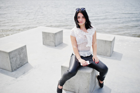 Portrait of brunette girl listening music at headphones from mobile phone, wear on women's leather pants and white blouse, sunglasses, against stone cubes at beach of lake.の写真素材