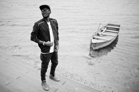 Portrait of stylish african american man on sportswear, cap and glasses against lake with boat....