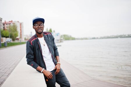 Portrait of stylish african american man on sportswear, cap and glasses against lake. Black men model fashion.の写真素材