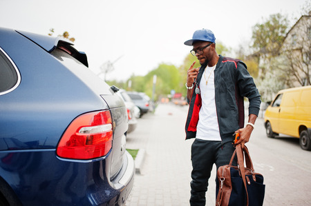 Portrait of stylish african american man on sportswear, cap and glasses walking with handbag and open car trunk. Black men model street fashion.の写真素材