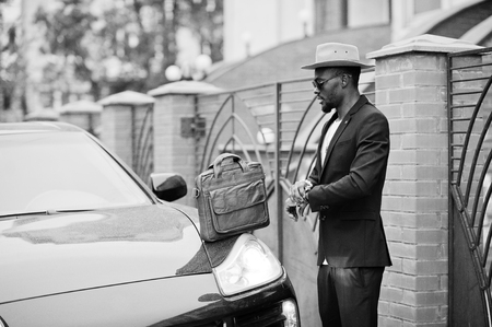 Stylish black man at glasses with hat, wear on suit with handbag against luxury car. Rich african american businessman. Look at watches.の写真素材