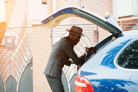 Stylish black man at glasses with hat, wear on suit with handbag against luxury car. Rich african american businessman.の写真素材
