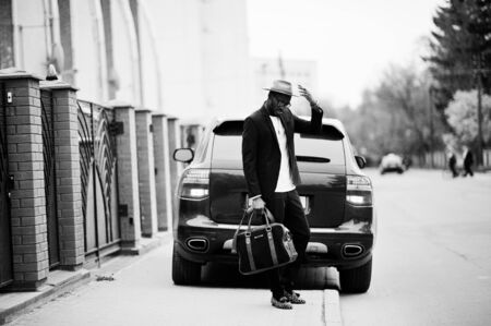 Stylish black man at glasses with hat, wear on suit with handbag against luxury car. Rich african american businessman.の写真素材