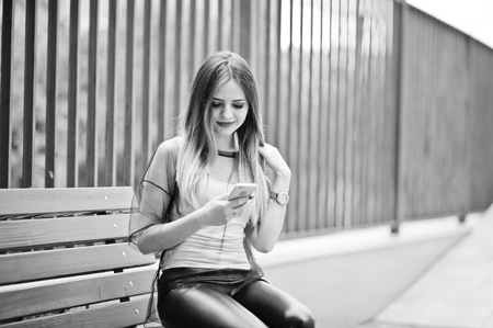Fashionable woman look at white shirt, black transparent clothes, leather pants, posing at street and sitting on bench with cell phone. Concept of fashion girl.の写真素材