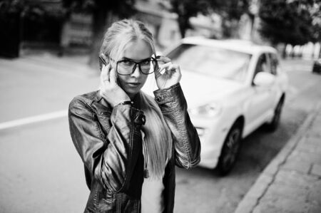 Stylish blonde woman wear at jeans, glasses and leather jacket with mobile phone, against luxury car. Fashion urban model portrait.の写真素材
