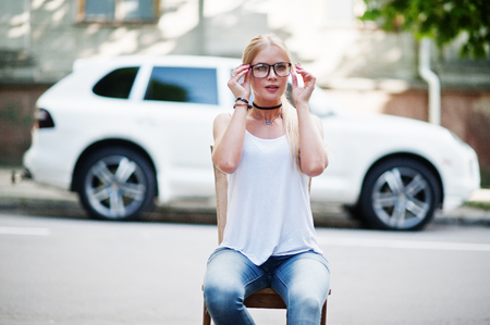 Stylish blonde woman wear at jeans, glasses, choker and white shirt near chair against luxury car. Fashion urban model portrait.の写真素材