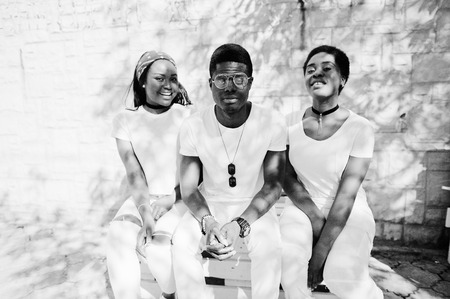 Three stylish african american friends, wear on white clothes sitting on bench. Street fashion of young black people. Black man with two african girls. の写真素材