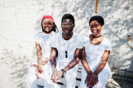 Three stylish african american friends, wear on white clothes sitting on bench. Street fashion of young black people. Black man with two african girls. の写真素材