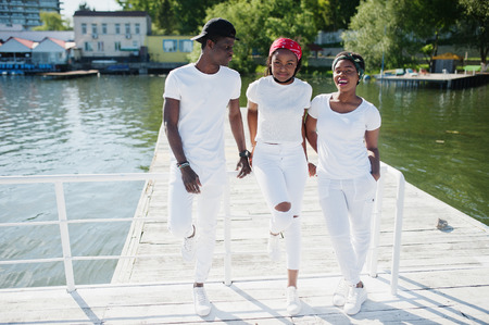 Three stylish african american friends, wear on white clothes at pier on beach. Street fashion of...