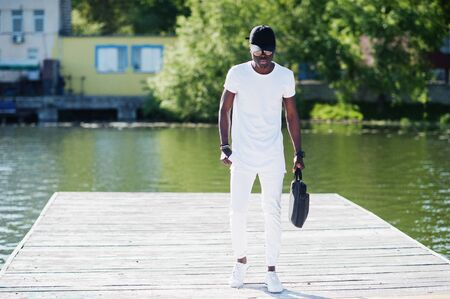 Portrait of stylish african american boy, wear on white clothes, glasses and cap with bag on hand against pier of lake. Street fashion of young black people.の写真素材