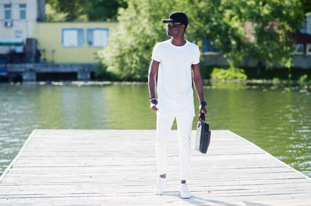 Portrait of stylish african american boy, wear on white clothes, glasses and cap with bag on hand against pier of lake. Street fashion of young black people.の写真素材