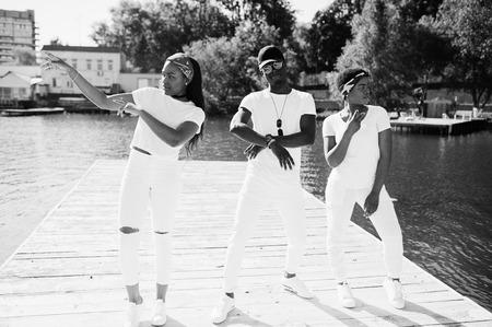 Three stylish african american friends, wear on white clothes at pier on beach showing dab. Street fashion of young black people. Black man with two african girls.の写真素材