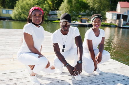 Three stylish african american friends, wear on white clothes at pier on beach. Street fashion of young black people. Black man with two african girls.の写真素材