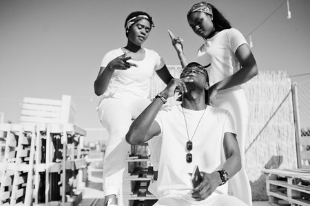 Three stylish african american friends, wear on white clothes at pier on beach. Street fashion of young black people. Black man with two african girls.の写真素材