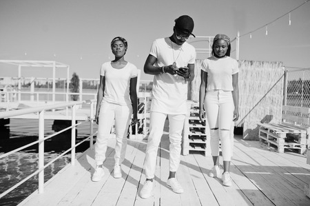 Three stylish african american friends, wear on white clothes at pier on beach. Street fashion of young black people. Black man with two african girls.の写真素材
