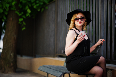 Blonde woman on black dress, sunglasses, necklaces and hat sitting on bench with mobile phone at hand and headphones.の写真素材