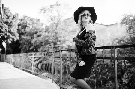Blonde woman on black dress, leather jacket, sunglasses, necklaces and hat against railings.の写真素材