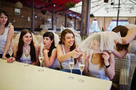 Group of cheerful girls at white shirts sitting at table and drink champagne on hen party.の写真素材