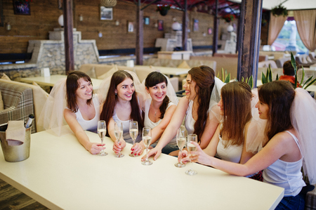 Group of cheerful girls at white shirts sitting at table and drink champagne on hen party.の写真素材