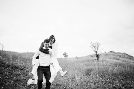 Happy bride riding her husband's back on their wedding day on the of hill with tall grass.の写真素材