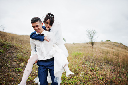 Happy bride riding her husband's back on their wedding day on the of hill with tall grass.の写真素材
