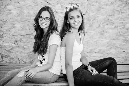 Two gorgeous girls sitting on wooden bars against veneer wall. Black and white photo.