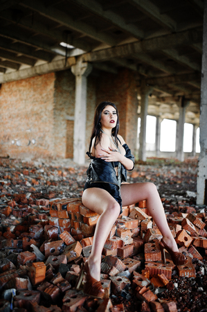 Brunette plus size sexy woman, wear at black leather jacket, lace panties, bra and high heels with chain at hands, siting on brick at abadoned place. BDSM theme.の写真素材