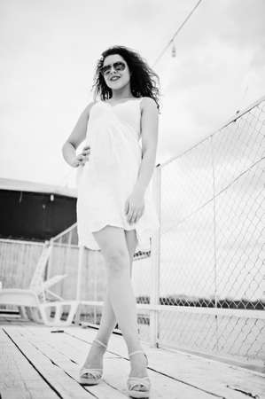Portrait of a stunning girl posing on a white wooden harbour next to the water with her marine white dress and sunglasses on. Black and white photo.の写真素材