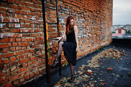 Red haired punk girl wear on black dress at the roof against brick wall with iron ladder.の写真素材
