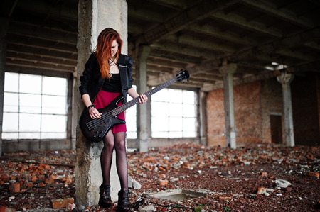 Red haired punk girl wear on black and red skirt, with bass guitar at abadoned place. Portrait of gothic woman musician.の写真素材