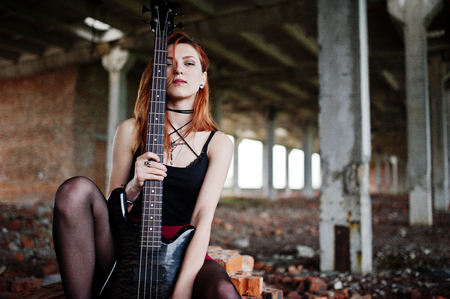 Red haired punk girl wear on black and red skirt, with bass guitar at abadoned place. Portrait of gothic woman musician.の写真素材