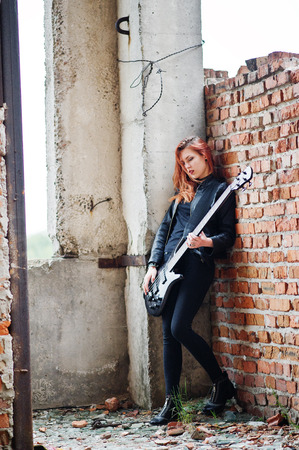 Red haired punk girl wear on black with bass guitar at abadoned place. Portrait of gothic woman musician.の写真素材