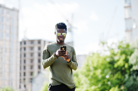 Attractive young african american guy wearing sunglasses uses his smartphone in the park with buildings in the background.の写真素材